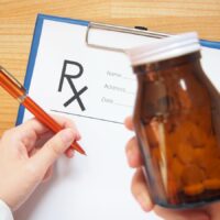 Female doctor hand holding blurred medical bottle and writing prescription form to patient at hospital or clinic. Pharmaceutical care, pain management and healthcare concept. Selective focus.