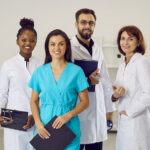 Multiethnic staff in clinic or hospital. Diverse team of happy doctors, general practitioners, specialists and nurses in scrubs and white lab coat uniforms standing in office and smiling at camera