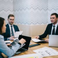 Blurred thoughtful adult female employee making report for directors in elegant suits while sitting at desk with documents and laptop during business meeting in office